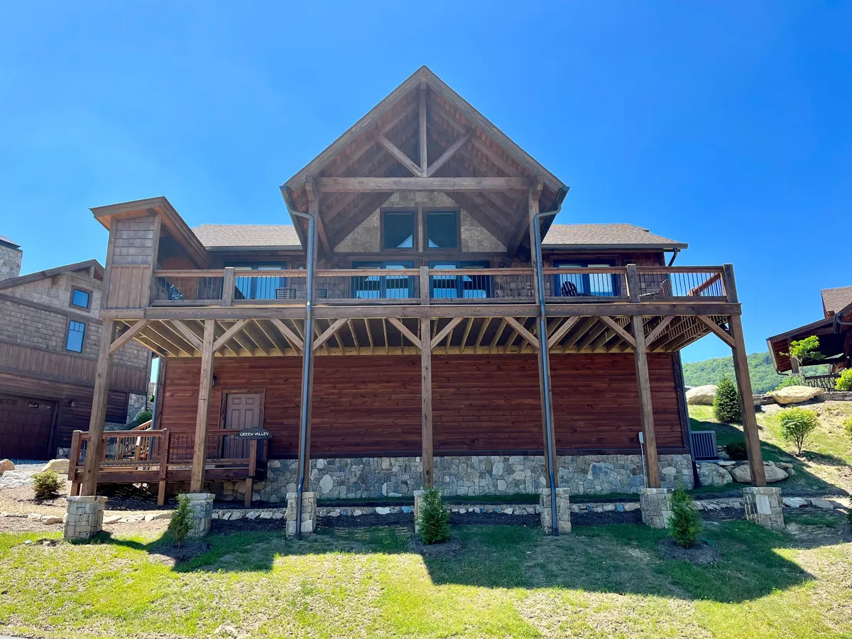 The image showcases a beautiful wooden mountain home with a spacious deck, stone accents, and sweeping views of rolling peaks and valleys. The deck overlooks a breathtaking landscape, perfect for relaxing or gathering, while the sunset bathes the scene in a warm, inviting glow.