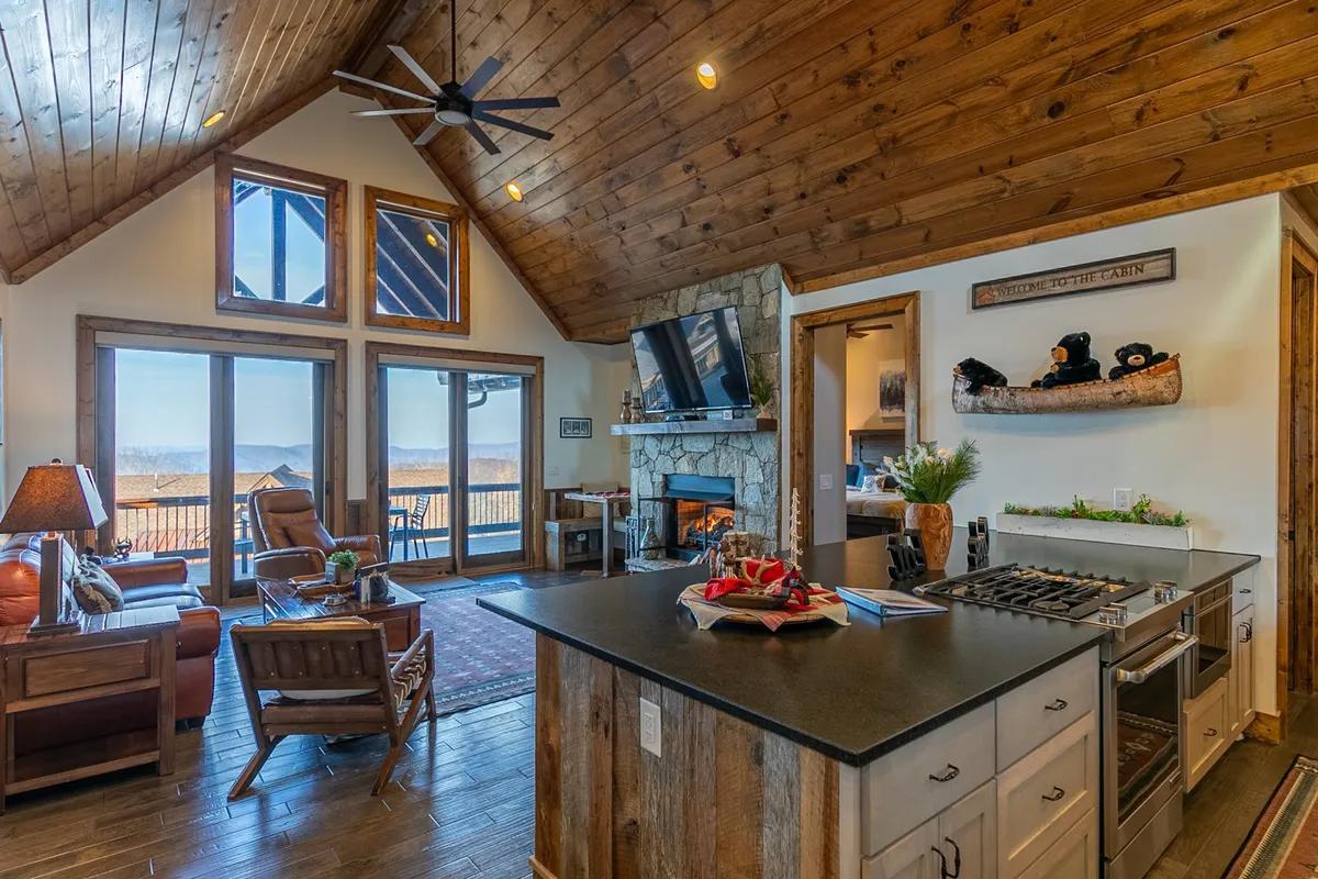 A spacious, open-concept living area featuring large windows that reveal panoramic mountain views. A modern stone fireplace anchors a comfortable seating area, while a sleek kitchen island with modern appliances is visible nearby—creating a seamless flow between relaxing and dining spaces.