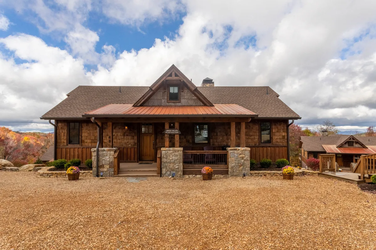 The exterior of a mountain-style home with a welcoming wooden front door, stone pillars, and a metal roof. Vibrant flower pots line the entrance, and guests are seen gathering on the porch, enjoying the surrounding natural beauty and mountain views.
