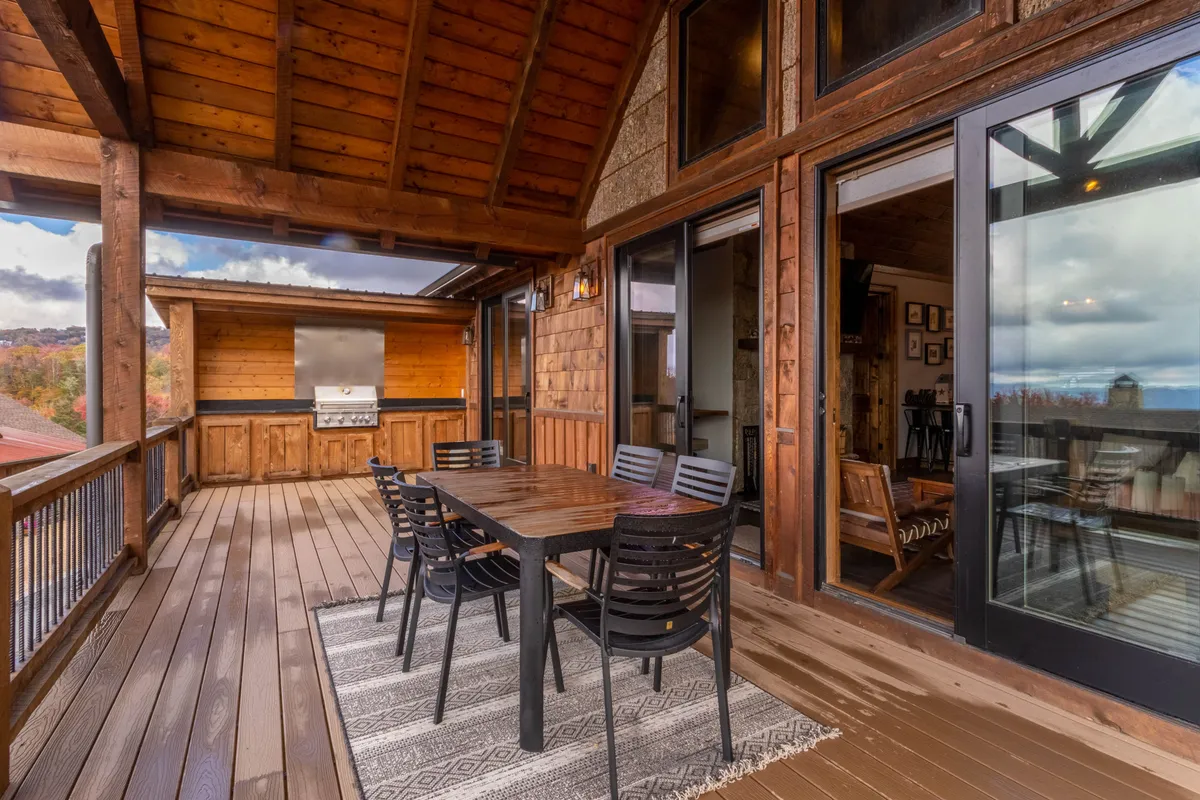 This image features a wooden dining table with black chairs situated on a spacious deck. A stone grill area is visible nearby, along with large glass doors that connect to the interior. The setup is inviting for outdoor dining, barbecuing, and gatherings, with the backdrop suggesting scenic mountain views.