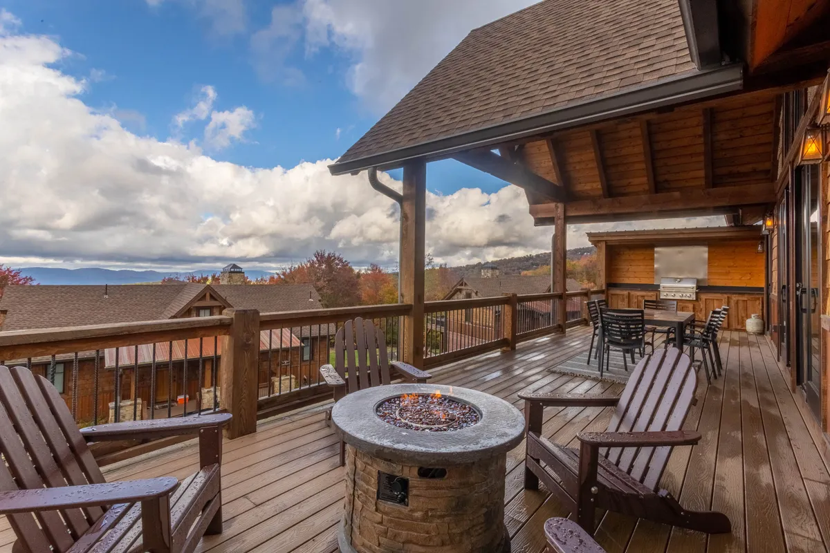 A cozy outdoor fire pit area is the focal point, surrounded by deck seating and set against a spectacular mountain view. The scene exudes comfort and sociability, ideal for gatherings around the fire as the sun sets.