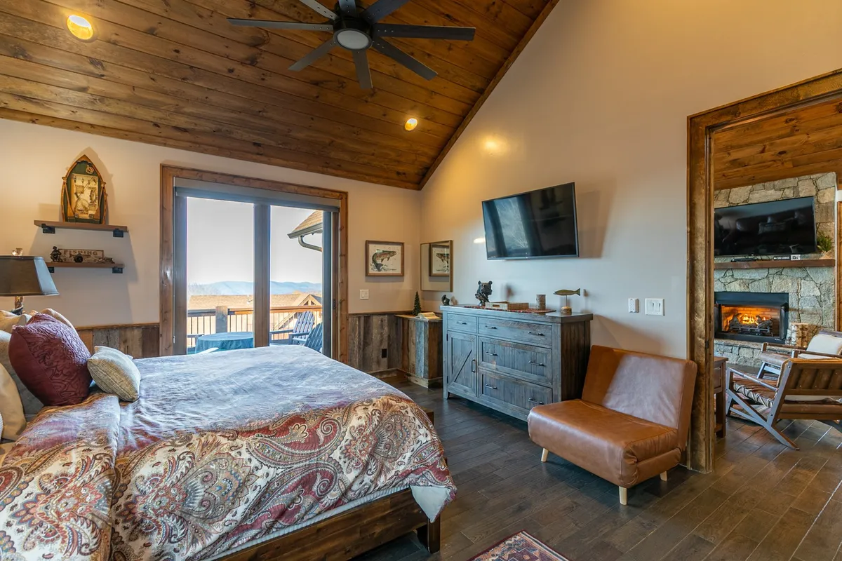 A spacious king bedroom featuring ornate bedding and a comfortable armchair placed near sliding glass doors that open to a private balcony. The room includes a cozy fireplace, offering both warmth and a romantic ambiance. Natural light pours in, highlighting the inviting textures and the tranquil mountain atmosphere visible through the glass doors.
