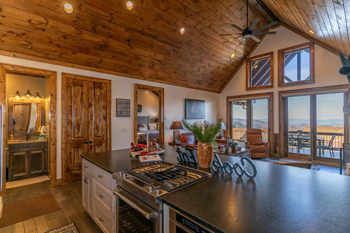 A bright, airy kitchen and lounge area with a large island, decorative plants, wooden accents, and oversized windows that flood the space with natural light and scenic views. Cozy seating invites guests to relax and socialize.