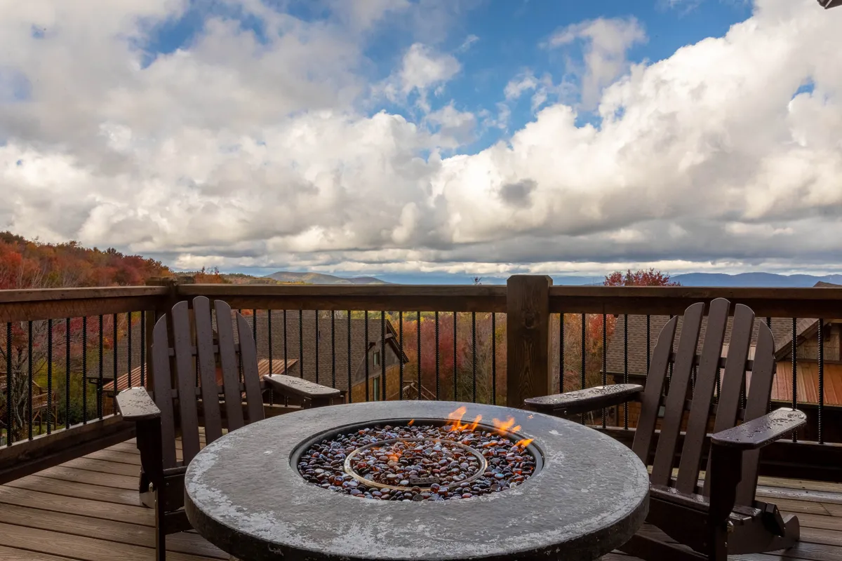 A tranquil outdoor setting featuring a circular stone fire pit surrounded by wooden chairs, all facing expansive mountain vistas under a cloudy sky. The layout encourages gathering, relaxation, and soaking in long-range views, with hints of lush greenery surrounding the area.