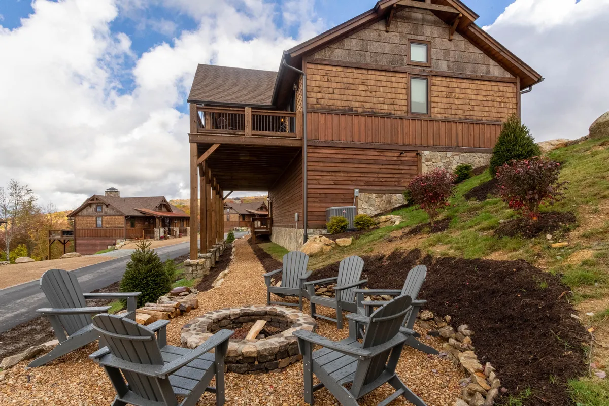 An inviting exterior scene with a fire pit surrounded by Adirondack chairs, set against the backdrop of a wooden house and lush natural surroundings. The area is designed for groups and families to gather, enjoy the outdoors, and share stories or meals around the fire.