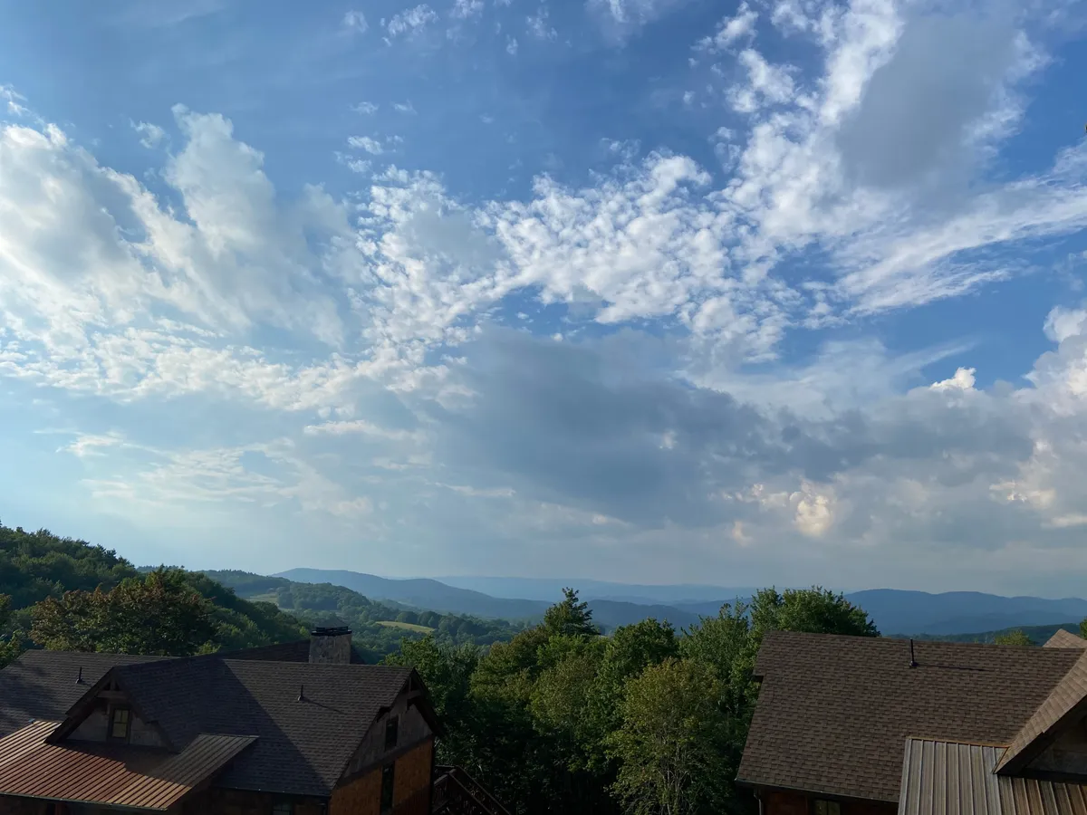 A panoramic view of a majestic mountain range under dramatic clouds, with the tree line and rooftops of nearby structures visible in the foreground. The scene hints at the vast outdoor possibilities, from hiking and picnicking to stargazing.